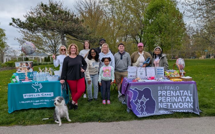 Community members and organization representatives gather behind Buffalo Prenatal Perinatal Network and Fidelis Care resource tables at an outdoor park event.