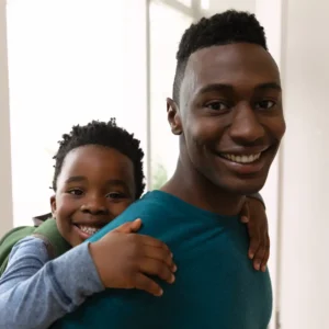 Adult and child smiling indoors, with the child riding on the adult’s back, both looking at the camera.